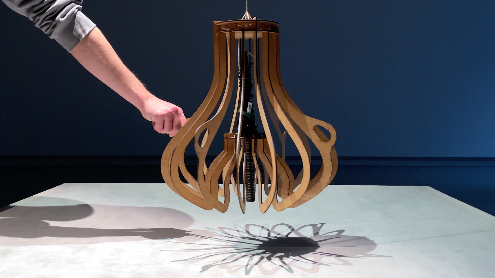 Echoes in Space — a hand swinging the wooden pendulum, casting a flower-shaped shadow